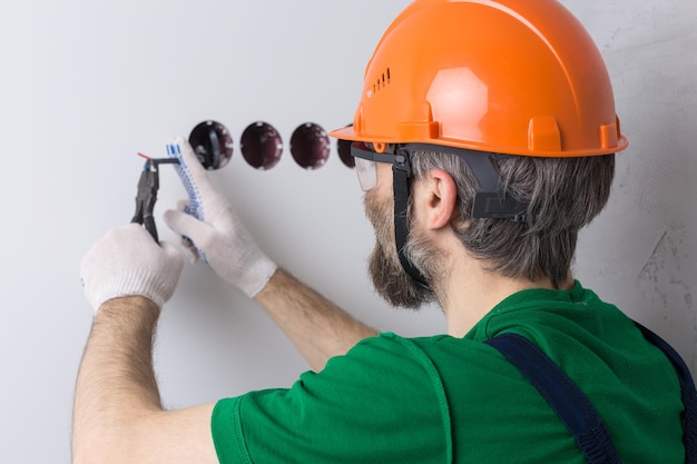 Ein Elektriker installiert Steckdosen in der Wohnung. Ein Typ mit orangefarbenem Helm und Overall macht Elektrik im Haus