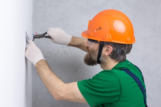 Ein Elektriker installiert Steckdosen in der Wohnung. Ein Typ mit orangefarbenem Helm und Overall macht Elektrik im Haus