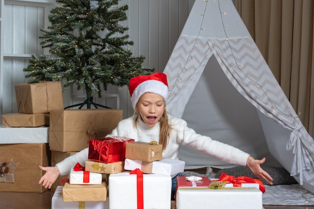 Ein emotionales Mädchen mit Weihnachtsmütze sitzt auf dem Boden neben einem Stapel ihrer Geschenke im Kinderzimmer Das Kind freut sich über Geschenke