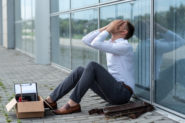 Ein entlassener büroangestellter sitzt auf dem boden in der nähe eines modernen bürogebäudes. der mann ist sehr besorgt über die entlassung. der arbeiter bedeckte sein gesicht mit den händen.