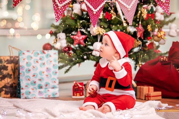 Ein entzückender kleiner Junge, der als Weihnachtsmann verkleidet ist, spielt in der Nähe eines geschmückten Weihnachtsbaums im Wohnzimmer.