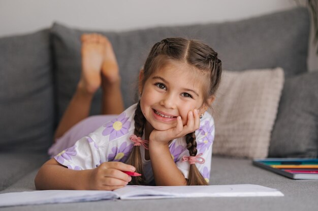 Ein entzückendes kleines Mädchen mit wunderschöner Zopffrisur liegt zu Hause auf dem Sofa und zeichnet ein Bild für