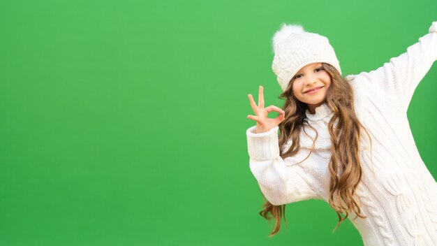 Ein entzückendes Model mit langen lockigen Haaren in einem warmen Winterpullover und einer Strickmütze schaut um die Ecke und zeigt eine Handbewegung OK. Schönes Baby mit lockigem Haar auf grünem Hintergrund.