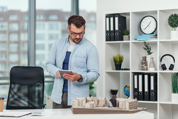 Ein erfahrener männlicher Architekt im Büro inspiziert das Projekt eines Wohnkomplexes und führt Berechnungen mit einem Tablet-PC durch.