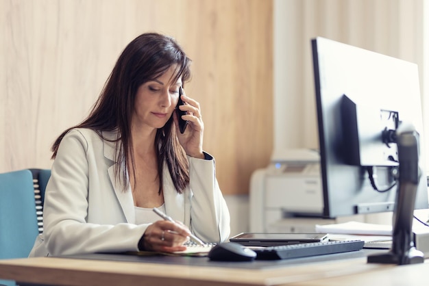 Ein erfahrener Manager im Anzug arbeitet an einem Computer im Büro und überprüft die Finanzergebnisse mit einem Kollegen per Handy