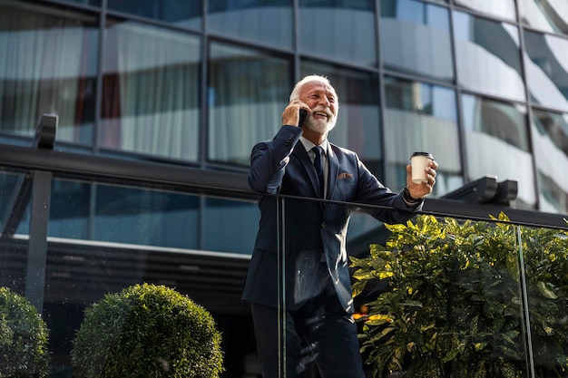 Ein erfolgreicher Senior CEO lehnt am Geländer, trinkt Kaffee und führt ein wichtiges Telefonat