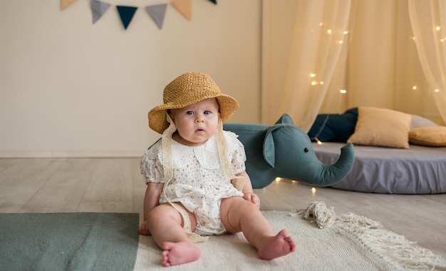 Ein ernstes kleines Mädchen in einem Bodysuit und einem Strohhut sitzt im Kinderzimmer auf dem Boden