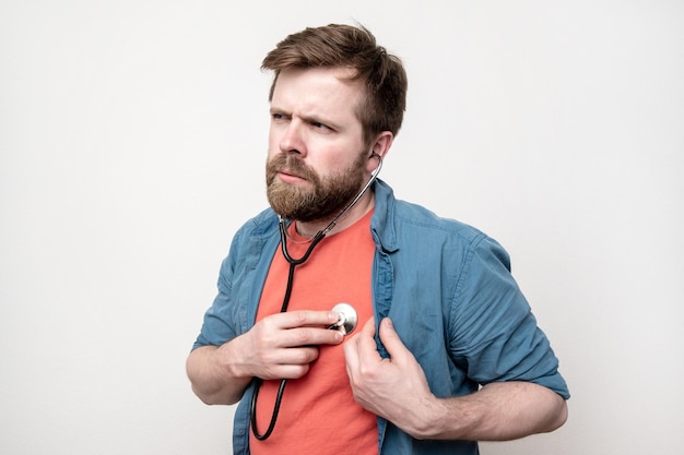 Ein ernsthafter bärtiger Mann hört mit einem Stethoskop auf sein Herz und schaut nachdenklich weg