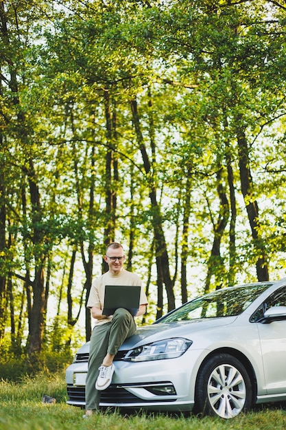 Ein ernsthafter Mann mit Brille arbeitet an einem Laptop auf der Motorhaube seines Autos Fernarbeit in der Natur Arbeiten an einem Laptop online Arbeiten an einem Computer während der Reise