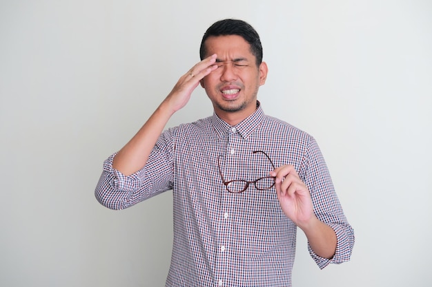 Ein erwachsener asiatischer Mann leidet unter Augenschmerzen, während er seine Brille hält