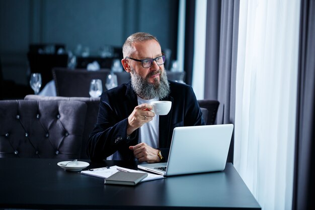 Ein erwachsener männlicher Geschäftsmann arbeitet an einem neuen Projekt und schaut sich die Aktiencharts an. Sitzt mit Kaffee am Tisch. Schaut auf den Laptop-Bildschirm und trinkt Kaffee.