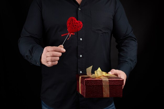 Ein erwachsener Mann in einem schwarzen Hemd hält eine rote Pappschachtel mit einer goldenen Schleife in der Hand