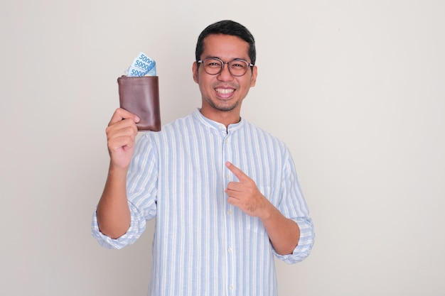 Ein erwachsener Mann lächelt glücklich und zeigt auf eine Brieftasche voller Geld, die er hält