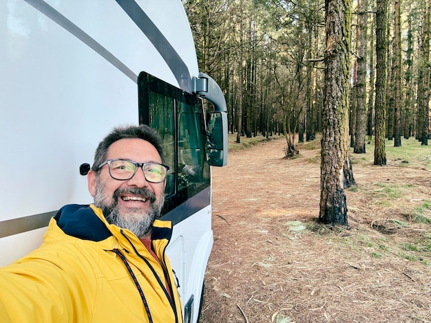 Ein erwachsener Mann macht ein Selfie vor einem Wohnwagen, der im Naturpark im Freien mit Bäumen parkt. Menschen und Reisen, alternativer Lebensstil, Wohnwagen, Urlaub, Mietfahrzeug, Freizeit.