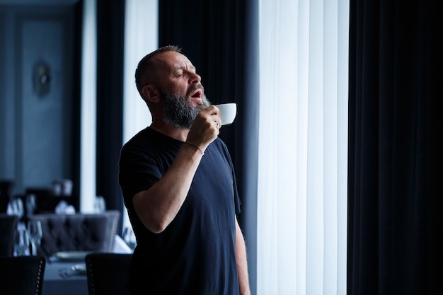 Ein erwachsener Mann mit Bart trinkt Kaffee und schaut aus dem Fenster. Erholungskonzept für den Arbeitstag