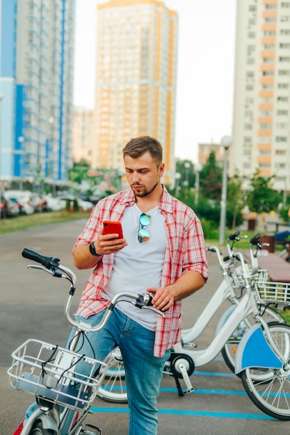 Ein erwachsener Mann mit Bart und Hemd steht mit einem Smartphone in der Hand vor dem Hintergrund einer Reihe von Mietfahrrädern und nutzt ein mobiles Programm zum Teilen von Fahrrädern