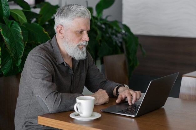 Ein erwachsener Mann mit grauen Haaren und Bart arbeitet an seinem Laptop in seinem Büro oder Wartebereich am Flughafen