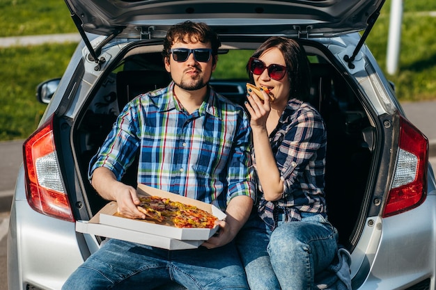 Ein erwachsenes, stilvolles Paar sitzt am Kofferraum eines Autos und isst einen Pizzasnack im Freien