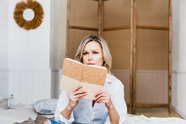 Ein europäisch aussehendes Mädchen sitzt auf einer Matratze und trägt eine weiße Bluse, Jeans, Mode, Schönheit, Stil, Tagebuch, Notizblock., Schreibt, schreibt