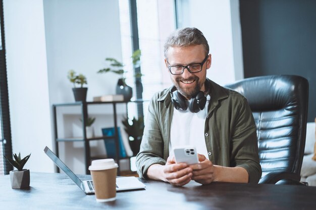 Ein europäischer Mann macht im Büro eine Pause und nutzt das Telefon, um Textnachrichten zu senden, die das Konzept einer einfachen Kommunikation veranschaulichen. Lässige und entspannte Arbeitsumgebung