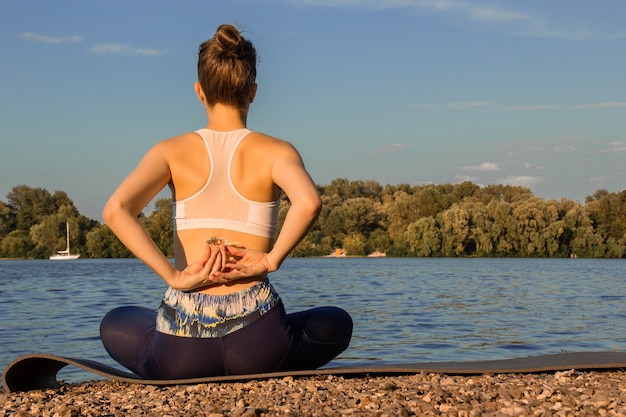 Ein europäisches mädchen in einem leichten trainingsanzug praktiziert yoga am flussufer auf einer gummi-yogamatte