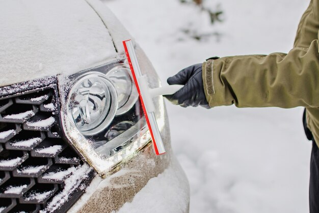 Foto ein fahrer, der autolichter im wintertag reinigt. schneebedecktes auto bei kaltem wetter. glasreinigungswerkzeug. scheinwerfer eines autos aus schnee im winter. bereiten sie ein auto für eine reise im winter vor.