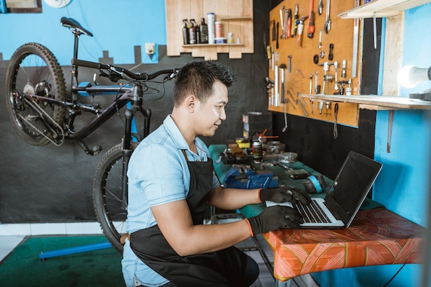 Ein Fahrradmechaniker in einer Schürze mit Handschuhen sitzt, während er mit einem Laptop nach Ersatzteilen sucht