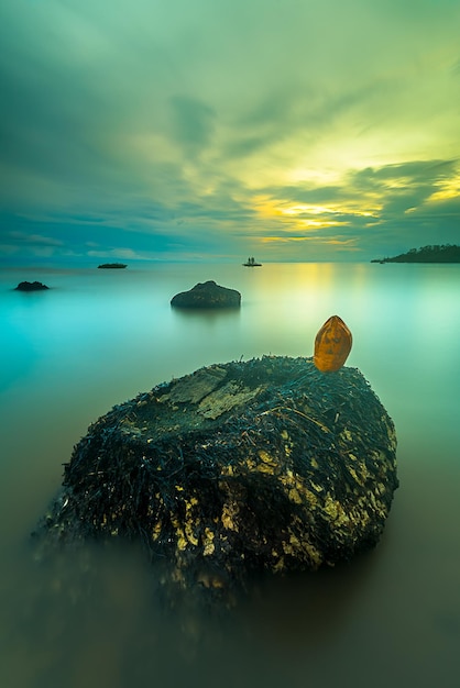 Ein Felsen im Wasser mit blauem Himmel und der untergehenden Sonne.