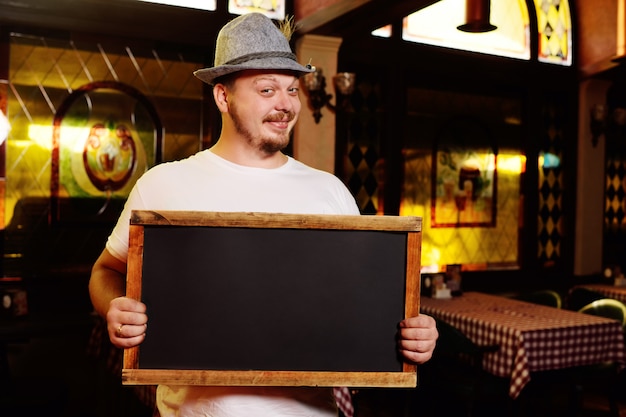 Ein fetter netter Mann in einem bayerischen Hut mit einer Feder während der Feier des Oktoberfestes hält ein Zeichen oder eine Tafel in seinen Händen