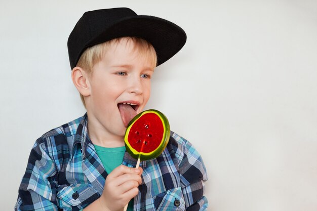 Ein Foto des lustigen kleinen Jungen in der trendigen Mütze und im Hemd, die riesigen Lutscher halten und lecken, der mit Glück beiseite schaut