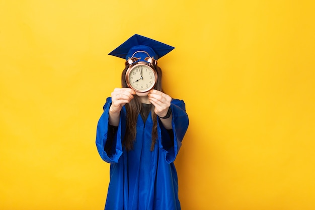 Ein Foto einer Studentin, die gerade ihren Abschluss gemacht hat und eine kleine Uhr in der Schrift ihres Gesichts in der Nähe einer gelben Wand hält