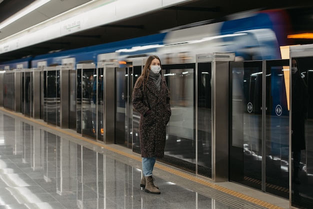 Ein Foto in voller Länge einer Frau in einer medizinischen Gesichtsmaske, um die Ausbreitung des Coronavirus zu vermeiden, die auf dem U-Bahnsteig auf einen ankommenden Zug wartet. Mädchen mit Mundschutz hält soziale Distanz