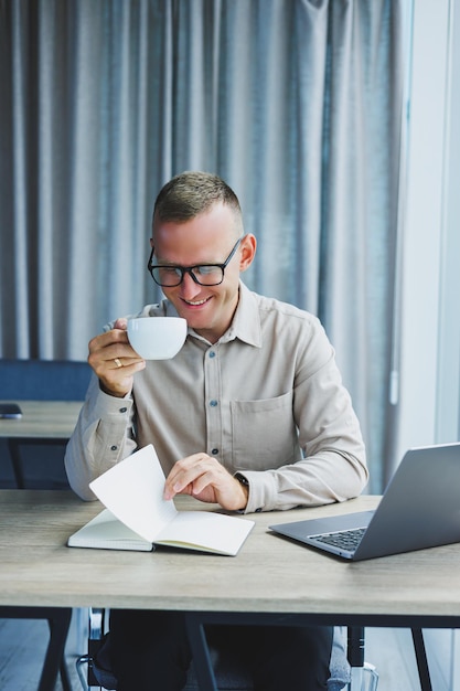 Ein Freiberufler mit Brille arbeitet an einem Laptop und trinkt Kaffee. Ein Manager sitzt an einem Tisch im Büro und arbeitet an einem Laptop. Freiberuflicher Arbeitsplatzmitarbeiter bei Fernarbeit