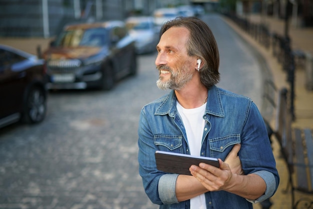 Ein freiberuflicher Mann mittleren Alters nutzt GPS oder ruft ein digitales Tablet an, während er durch die Straßen der Stadt geht. Reifer Mann nutzt drahtlose Kopfhörer, um zu reden oder Musik zu hören, während er durch die Straßen der Altstadt fährt