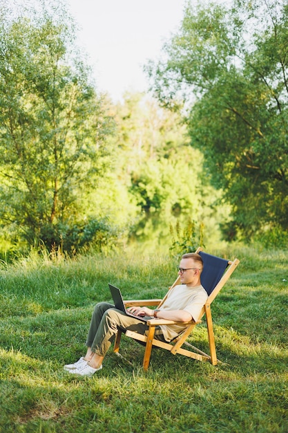 Ein freiberuflicher Mann sitzt auf einem Holzstuhl in der Natur und arbeitet online auf einem Laptop. Ein Mann reist und arbeitet aus der Ferne auf einem Laptop. Büroarbeit in der Natur. Urlaub