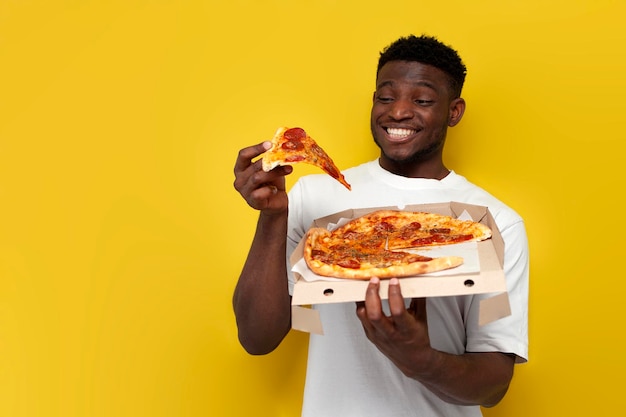 Ein freudiger afrikanisch-amerikanischer Mann in weißem T-Shirt hält eine Kiste Pizza und lächelt auf gelben Hintergrund