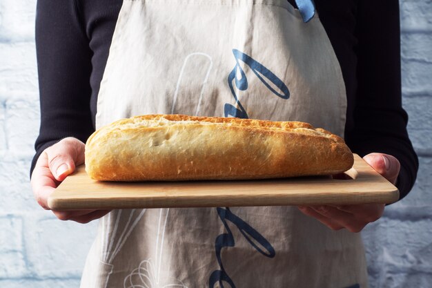Ein frisches Baguette Brot auf einem Holzbrett wird in den Händen einer Köchin gehalten.