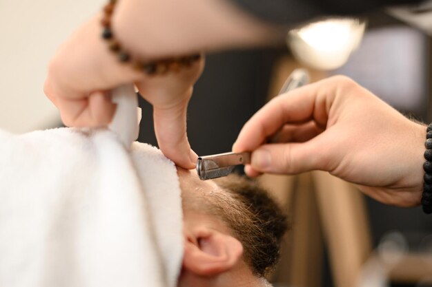 Ein Friseur rasiert die Wange eines bärtigen Kunden mit einem gefährlichen Rasiermesser und rasiert die Kontur des Bartes für die richtige Form.