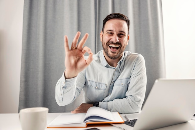 Ein fröhlich lachender Unternehmer in die Kamera und mit der Hand auf sein Home Office gestikulierend