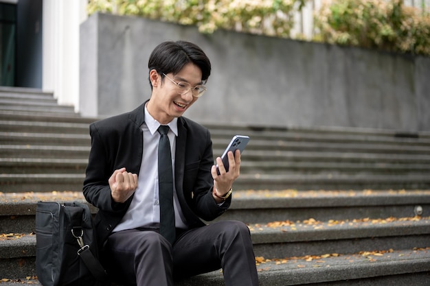 Ein fröhlicher asiatischer Geschäftsmann zeigt seine Faust und schaut mit einem glücklichen Gesicht auf sein Telefon