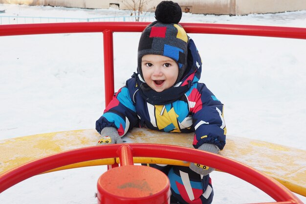 Ein fröhlicher Junge von 2 Jahren in Hut und Overall dreht sich im Winter auf einem Straßenkarussell. Freudig und fröhlich.