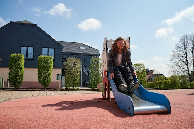 Ein fröhlicher junger kaukasischer Mann, der auf Kinderrutschen auf dem Spielplatz sitzt