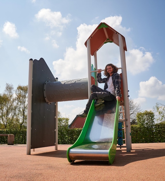 Foto ein fröhlicher junger kaukasischer mann, der auf kinderrutschen auf dem spielplatz sitzt