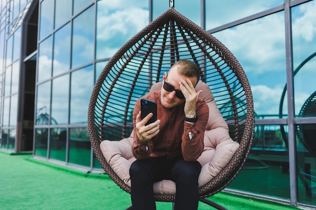 Ein fröhlicher junger mann in einem hemd mit einem telefon sitzt auf der terrasse eines bürogebäudes nach der arbeit nutzt das drahtlose internet ein positiver männlicher mitarbeiter mit brille spricht am telefon