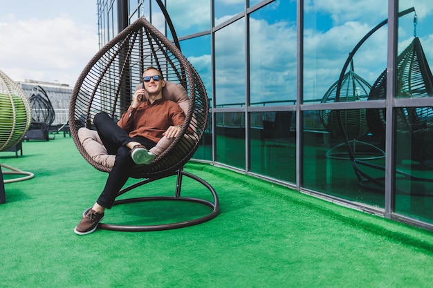 Ein fröhlicher junger mann in einem hemd mit einem telefon sitzt auf der terrasse eines bürogebäudes nach der arbeit nutzt das drahtlose internet ein positiver männlicher mitarbeiter mit brille spricht am telefon