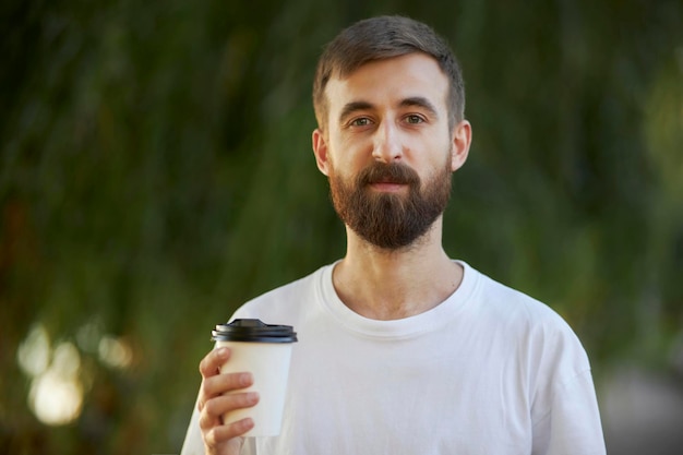 Ein fröhlicher Mann in einem weißen T-Shirt trinkt Kaffee aus einem weißen Pappbecher. Ein weißer Pappbecher