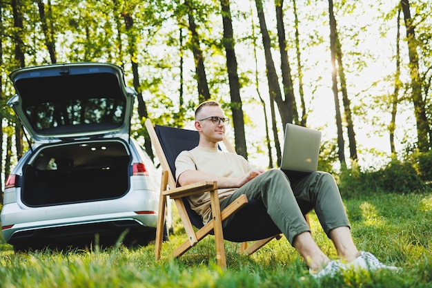 Ein fröhlicher Mann mit Brille sitzt auf einem Stuhl in der Natur und arbeitet im Urlaub online auf einem tragbaren Laptop. Freiberuflich tätig