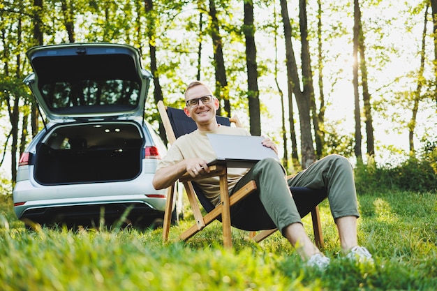 Ein fröhlicher Mann mit Brille sitzt auf einem Stuhl in der Natur und arbeitet im Urlaub online auf einem tragbaren Laptop. Freiberuflich tätig