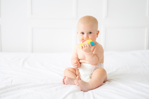 Ein fröhliches Baby in einer Windel sitzt auf einem Bett, das Kind hält ein Spielzeug und schaut in die Kamera