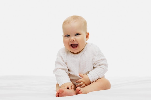 Foto ein fröhliches baby mit glänzenden blauen augen und einem freudigen lächeln sitzt auf einem weißen bett, gekleidet in ein behagliches weiß.